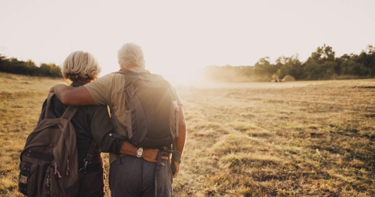 The backs of an older man and woman with arms around each other hiking into the sunset.