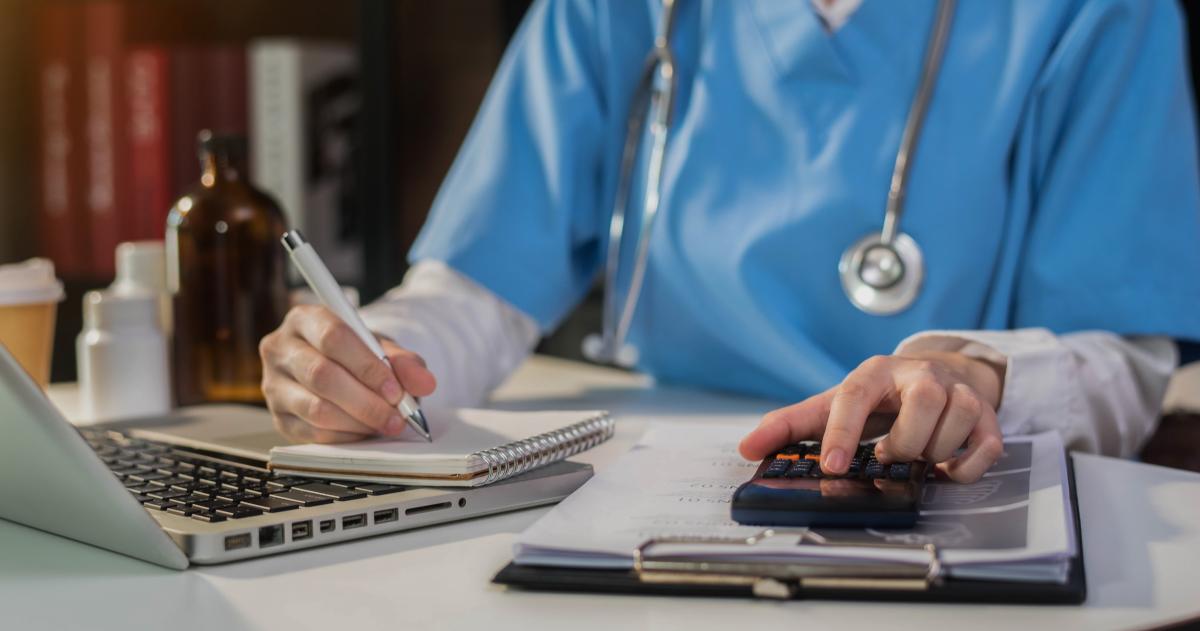 Person in scrubs with stethoscope using a calculator, notebook and laptop